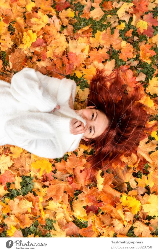 Beautiful redheaded woman lying on autumn leaves. Young woman smiling in autumn scenery 20s November October above view adult alone autumn fashion