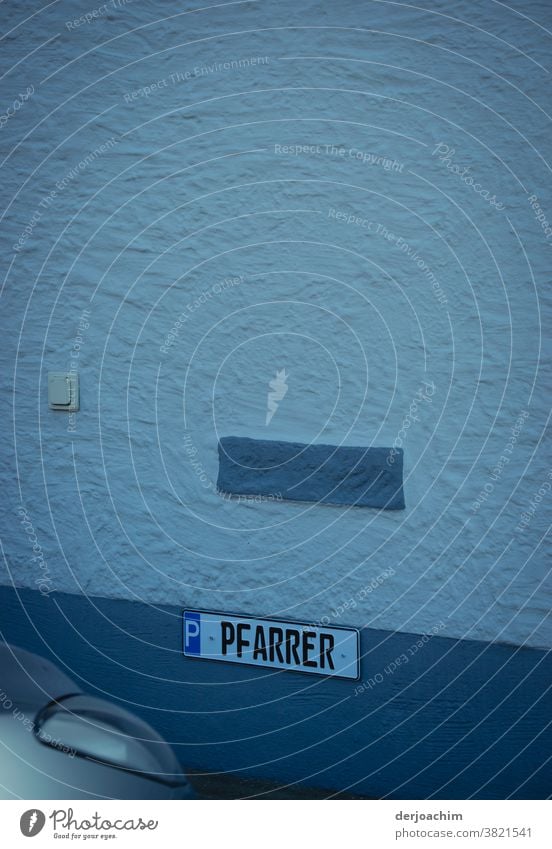 Parking for the PFARRER. Unfortunately it is occupied by an unknown car. The sign is attached to a white wall with blue gradations. In the lower edge of the picture on the left, a headlight is to be recognized.