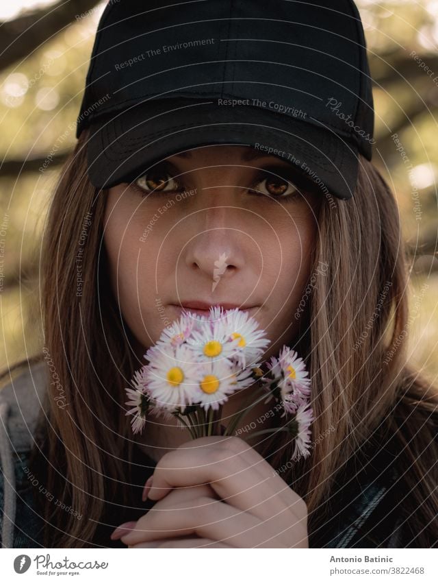 Closeup of an attractive brunette holding a bouqet of small white hand picked flowers on her lips, bright colorful amber eyes looking straight into the camera. Wearing a black cap in nature