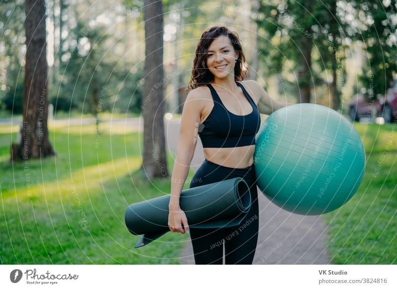 Sporty European woman in cropped top and leggings, carries rolled up karemat and fitness ball, going to have aerobics exercises, poses outdoor against green nature background, has pilattes class