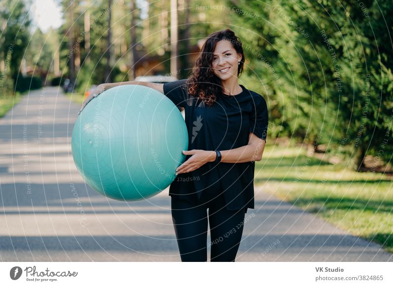 Outdoor shot of active brunette woman exercises with fitness ball poses at  green grass, dressed in active wear, enjoys sunny day and fresh air in  park, keeps fit, makes gymnastics exercises. 