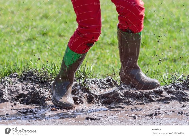 Legs in red trousers and green rubber boots jumping in a muddy puddle, so that the mud flies up Puddle slush Rubber boots Jump Muding Flying Inject Boots Joy