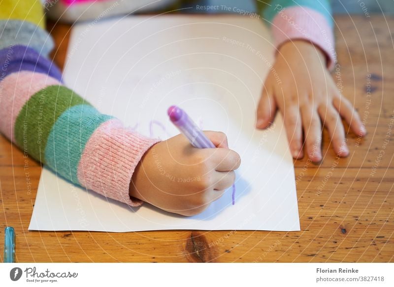 4 year old blonde girl with a brightly colored striped sweater sits at a wooden table and draws with a purple pencil on a piece of white paper