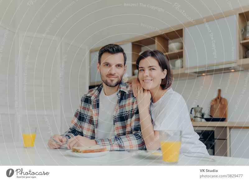 Positive brunette woman leans at shoulder of her husband, pose together at kitchen, enjoy delicious breakfast, have happy mood, look directly at camera, have good relationships. Family concept