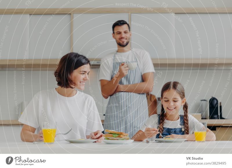 Happy mother talks to daughter while have breakfast. Father stands behind, prepared delicious dish for family. Friendly family members meet at kitchen during weekend, enjoy nice conversation