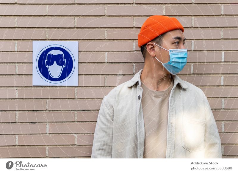 Asian man in mask standing near sing on building wall serious sign respirator obedience message symbol coronavirus new normal concentrate thoughtful pensive