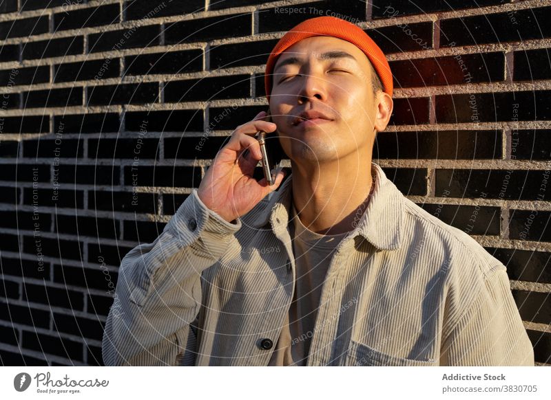 Dreamy Asian man taking on smartphone against brick wall content conversation phone call talk dreamy enjoy happy listen eyes closed device gadget modern
