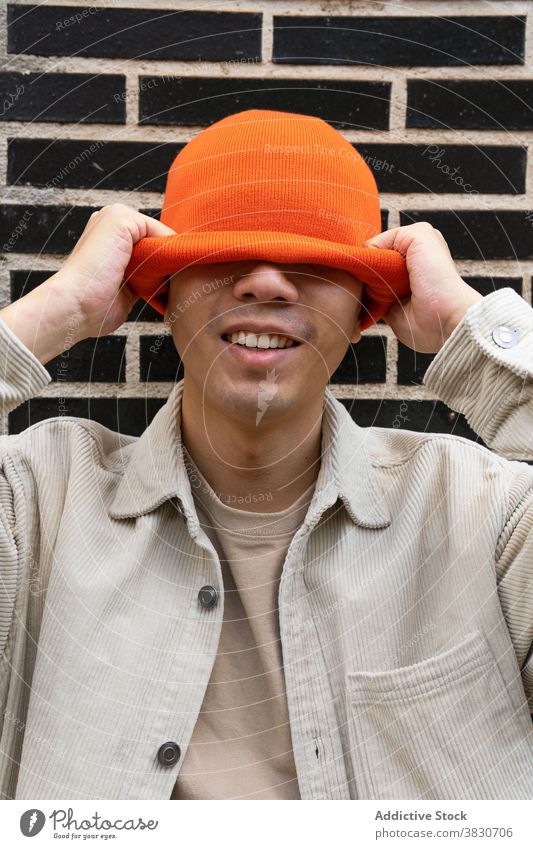 Positive man putting orange hat on head positive put on cover eyes happy headgear cool pull brick wall cheerful hipster fashion fun content pleasant glad