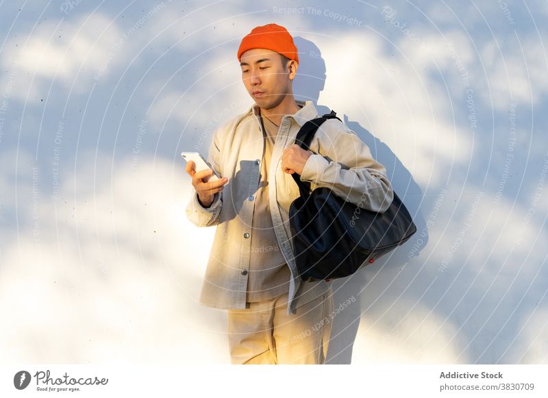 Asian man with travel bag using smartphone against white wall concentrate browsing cellphone focus surfing gadget modern young casual mobile calm device baggage
