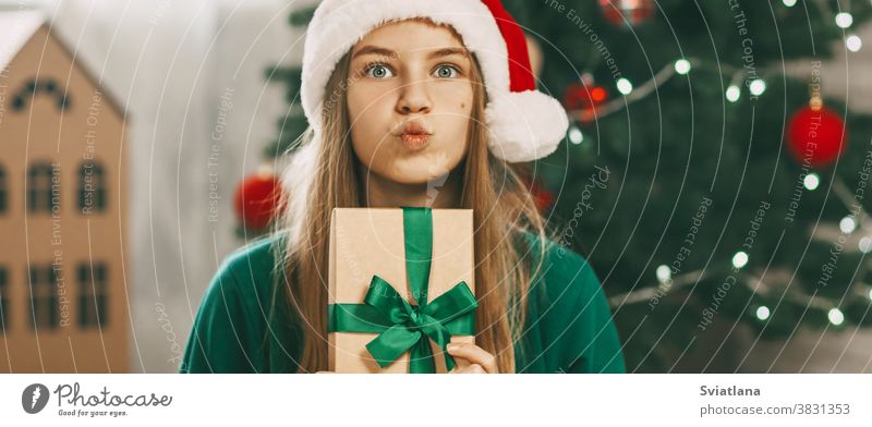 A beautiful girl with surprised eyes looks at the camera with a gift from kraft paper tied with a green ribbon. Christmas mood. Concept for New Years holiday at home.
