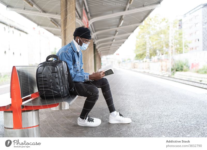 Ethnic man reading book on railway station wait train platform traveler mask coronavirus railroad male ethnic black african american trip new normal bench