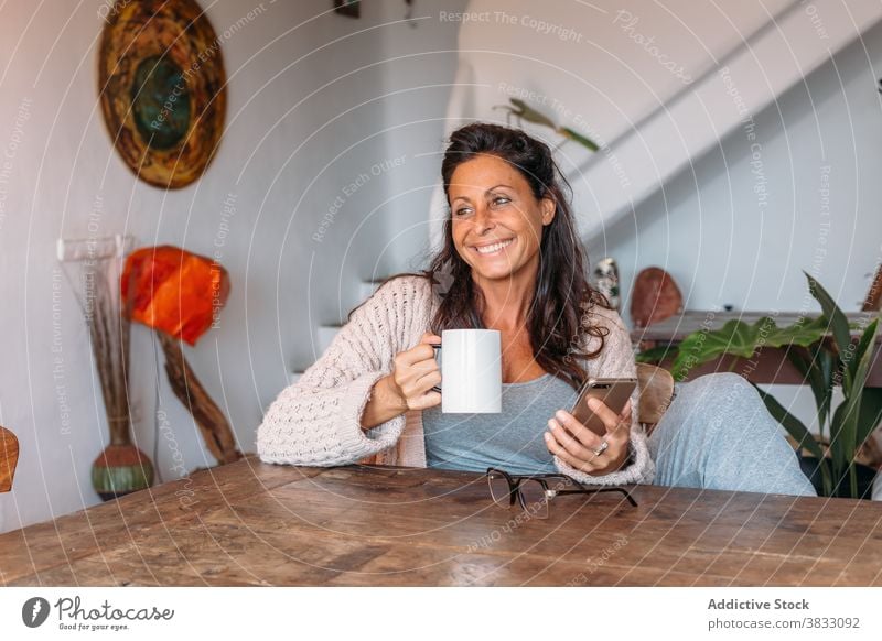 Happy woman with cup of coffee and smartphone sitting at table at home happy cheerful rest drink using adult ethnic female mobile device gadget beverage