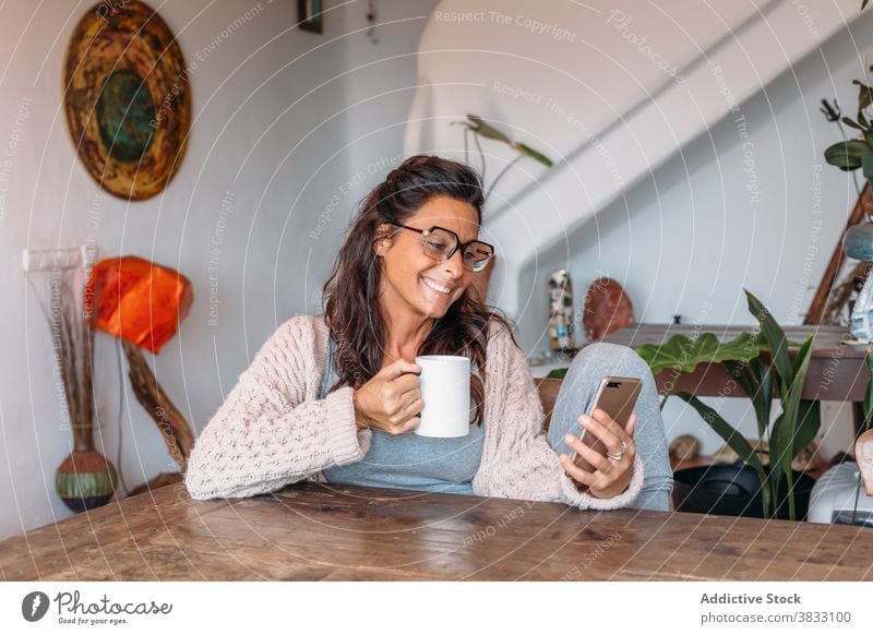 Positive ethnic woman with cup of coffee browsing smartphone at home happy cheerful rest drink using cozy comfort adult female mobile device gadget beverage