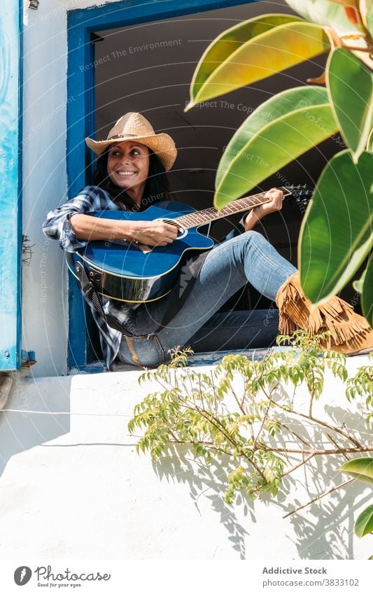 Happy woman in cowboy hat and boots playing guitar on windowsill country rural chill happy rest style cheerful ethnic female countryside smile lifestyle music