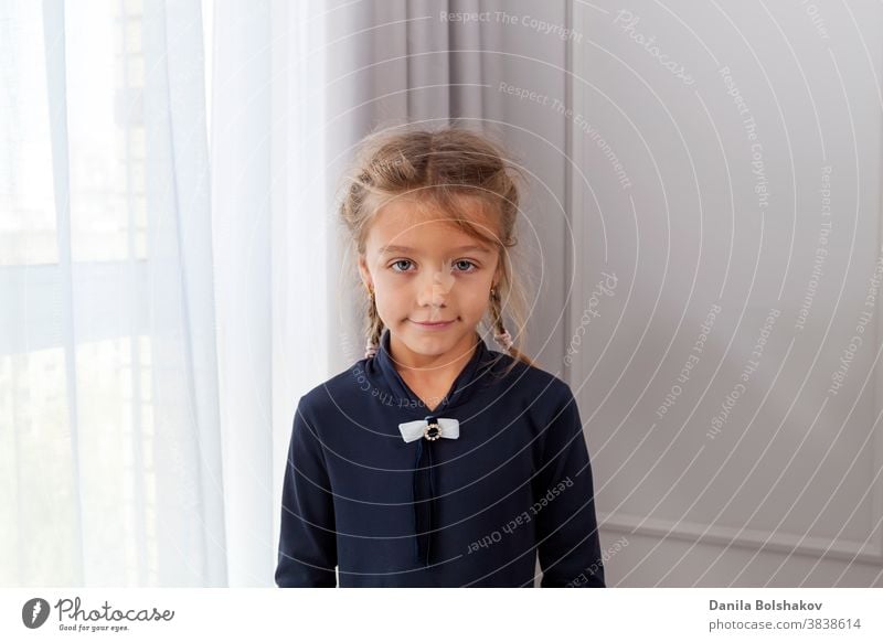 Close up shot of beautiful caucasian little girl with pigtails, looking at camera with charming smile, posing against light window and painted wall. Concept of happy childhood