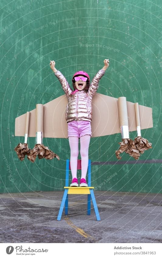 Cheerful child in carton jetpack rejoicing with raised arms wing plane play cardboard victory celebrate achieve success chair helmet triumph gesture cheerful