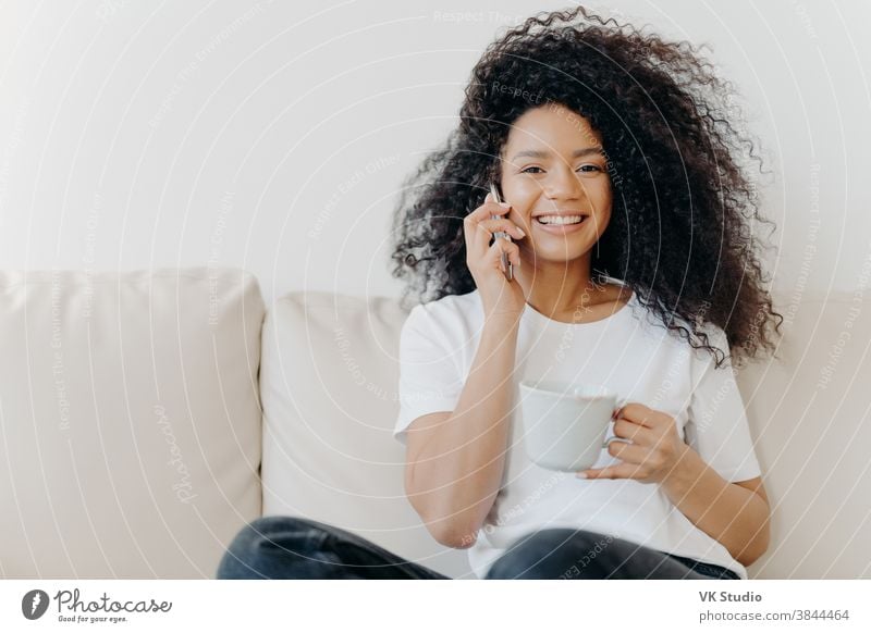 Horizontal shot of good looking woman has distant conversation via mobile phone, solves informal issues while talking, sits on sofa in living room, drinks tea, has broad smile, shows white teeth