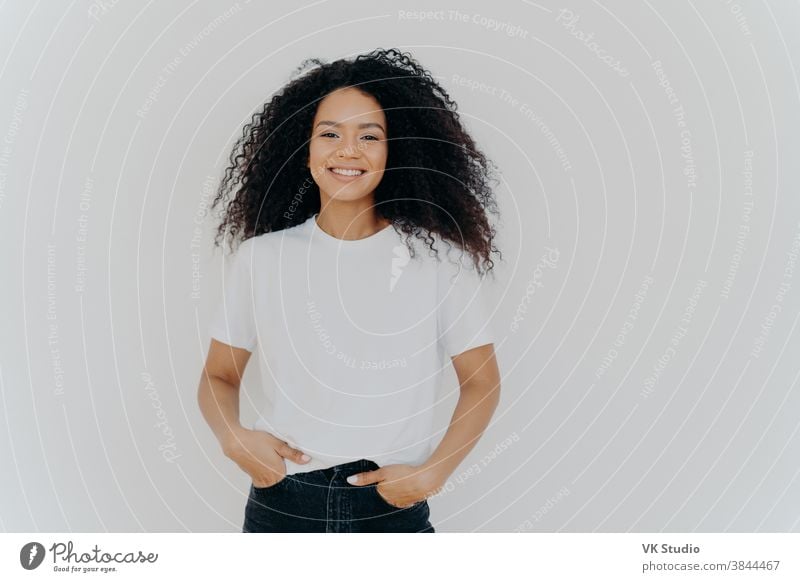 Isolated shot of young African American woman wears white t shirt, expresses good emotions, stands alone indoor, poses for photo, has casual talk with friend, enjoys free time. People and happiness