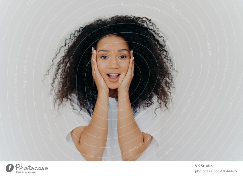 Photo of surprised cheerful woman with Afro haircut, keeps both hands on cheeks, has natural beauty, opens mouth, cannot believe in exciting news, wears t shirt, poses against white background.