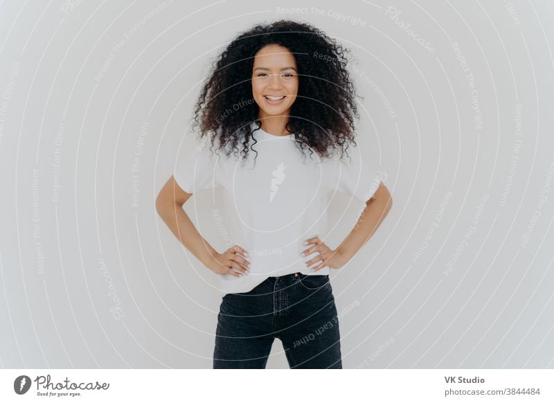 Photo of delighted curly woman keeps both hands on waist, smiles gently, has slim figure, wears white t shirt and black jeans, being in good mood, stands self assured against white background