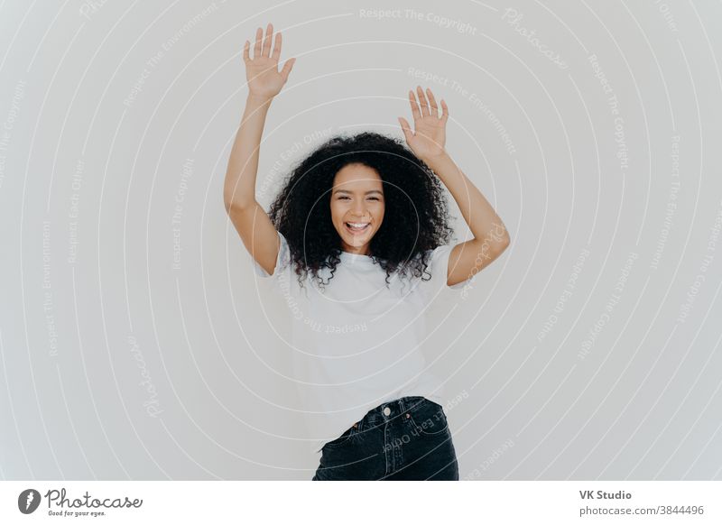 Half length shot of energetic carefree curly woman cheers with hands  raised, wears white t shirt and jeans, has lucky day, perfect weekend,  models against white background. Everybody put your hands up -
