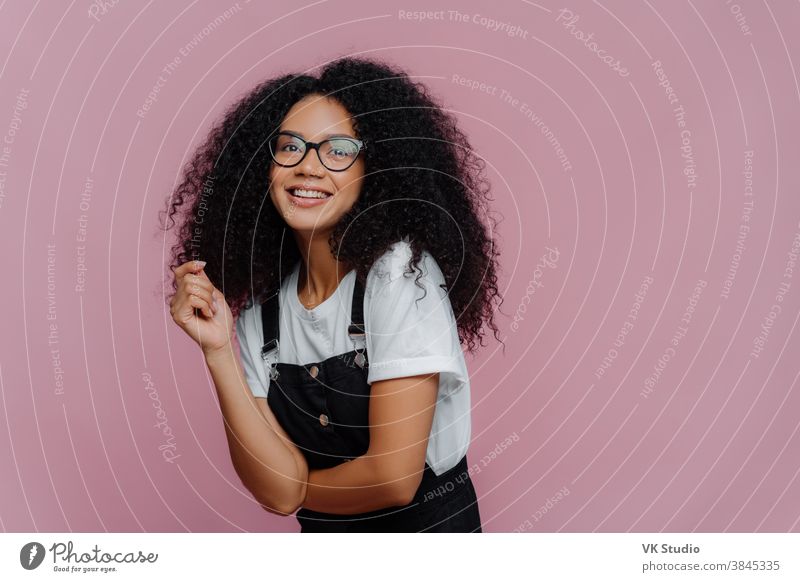 Studio shot of happy dark skinned lady with crisp hair, has dreamy expression, wears optical glasses, stylish clothes, imagines something pleasant, stands against bright violet background, free space