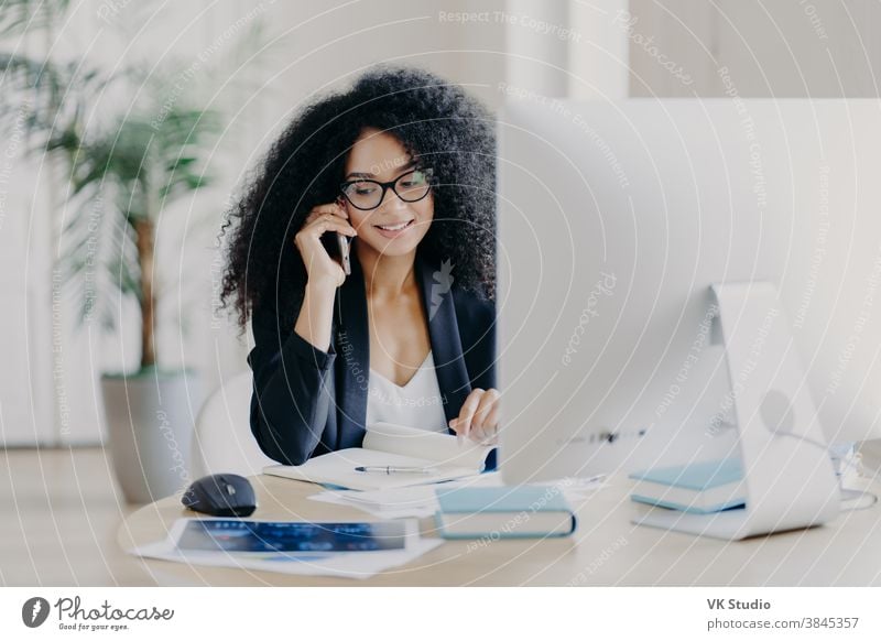 Photo of satisfied Afro American woman calls via smartphone, makes notes in notepad, involved in working process, sits at desk with computer, wears spectacles, formal clothes, has phone communication