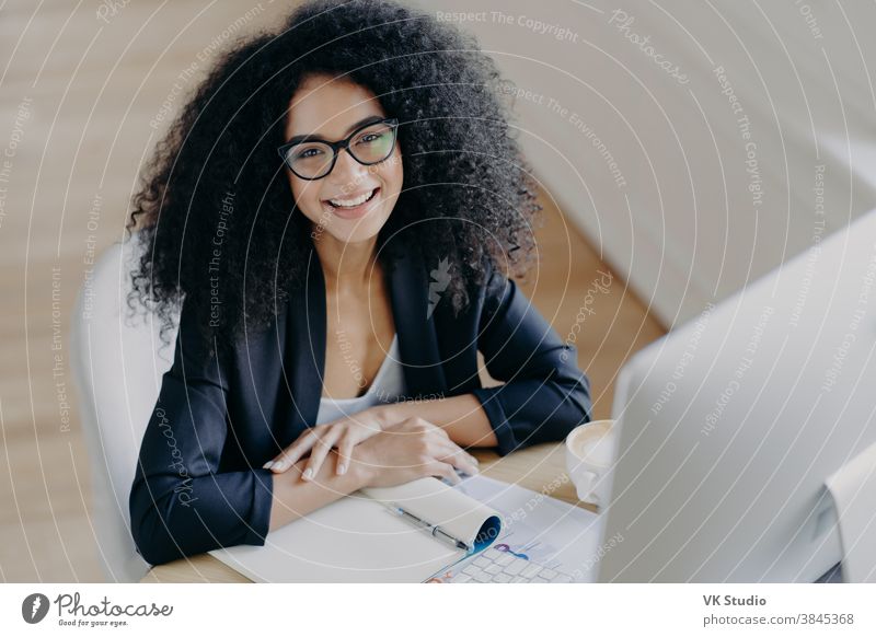 Top view of glad African American businesswoman smiles pleasantly, notes information, makes notes, wears transparent glasses, formal clothes, computer monitor in front, sits at desktop alone