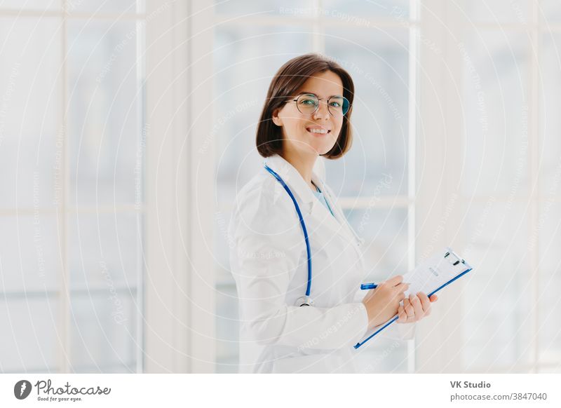 Horizontal shot of female doctor fills up medical form at clipboard, stands indoor, wears round glasses, white gown and stethoscope. General practitioner writes down notes, consults patients