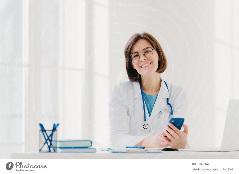Horizontal view of smiling professional doctor works in clinic, poses at modern hospital office with electronic gadgets, sends text messages on cellphone being at work. Health care, technology concept