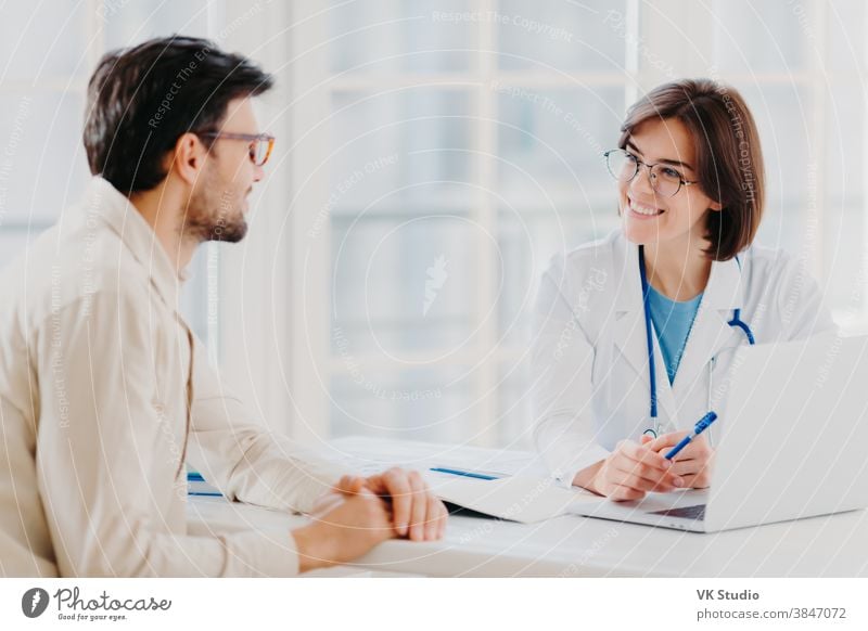 Skilled professional female general practitionist explains diagnosis and consults patient, demonstrates information in laptop, pose in clinical office, discuss scan results in diagnostic center