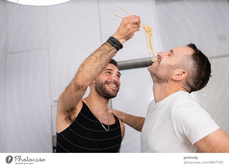 Man feeding pasta to his boyfriend in a fun way gay couple men homosexual spaghetti food cooking kitchen lgbt love lgbtq male relationship lovers people 30s
