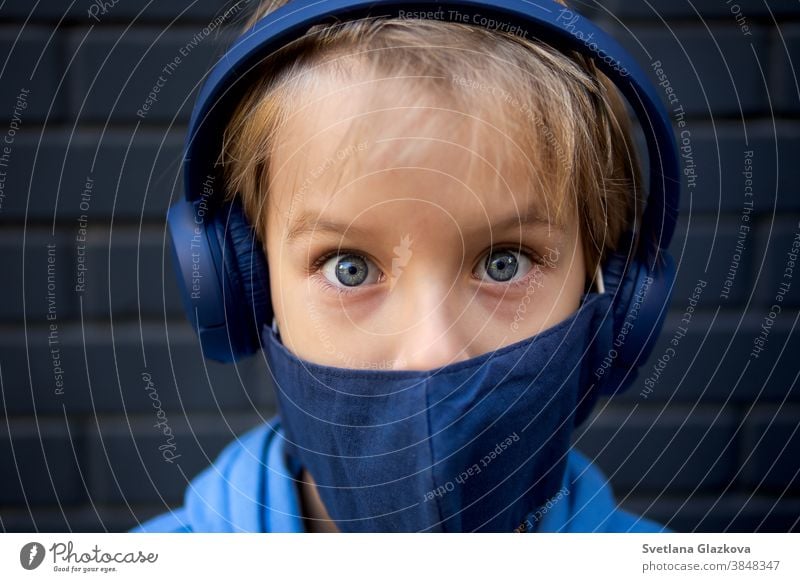 Close-up face portrait of a blonde caucasian boy. Looks at the camera. Dressed in a protective medical mask and blue headphones eyes close-up funny kid child