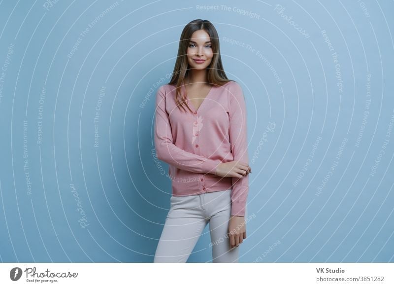 Fashionable brunette Caucasian woman dressed in pink jumper and white trousers, going to work, applies makeup, smiles pleasantly, isolated on blue background. People, style, lifestyle concept