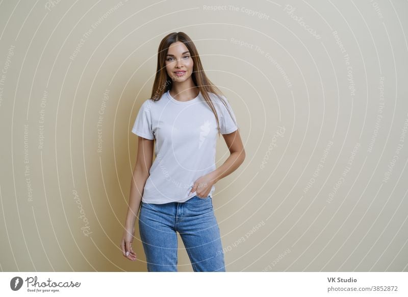 Studio shot of pretty slim woman keeps hand in pocket, wears casual t shirt and jeans, stands in relaxed pose, looks at camera self confident, isolated over beige background talks casually with client