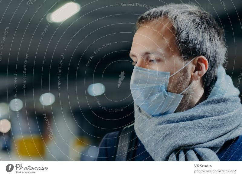 Pensive man wears protective mask against new coronavirus from China, wrapped scarf around neck, looks somewhere, thinks about epidemic situation. Influenza, flu symptoms, virus, treatment concept