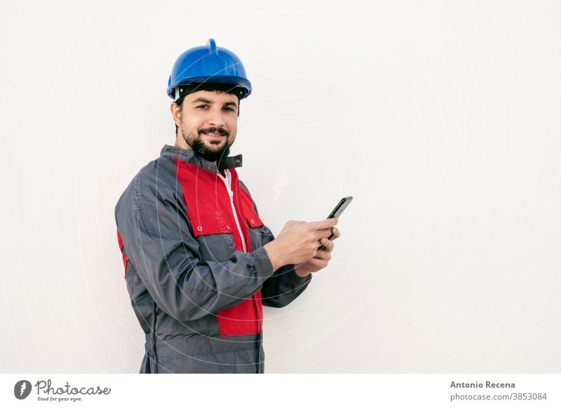 Bearded worker man with smart phone looking at camera smiling laborer uniform helmet pandemic virus white background wall hardhat protection goatee beard arab