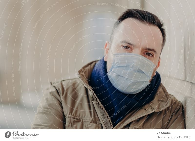 Outdoor shot of young European man has coronavirus symptoms, wears protective surgery mask, tries protect from viruses and infections, needs to see doctor. Street and air contamination problem