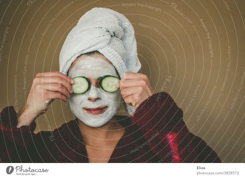 Woman with bathrobe and towel on her head enjoys a face mask and holds two slices of cucumber in front of her eyes. Wellness, face care. Face mask