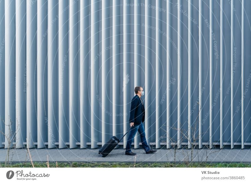 Horizontal panoramic shot of man passenger arrives in own country because of quarantine and pandemic situation in world, walks with suitcase, poses outdoor against metal fence, wears face mask