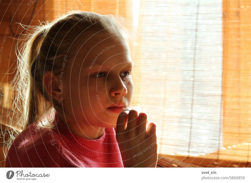 A blonde teenage girl sits by a window in the daylight through a curtain and looks forward thoughtfully, her hand to her chin. Portrait of a pensive blonde girl.