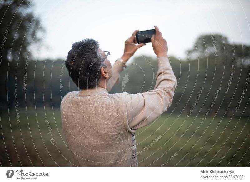 Back side portrait of an old Indian Bengali man in woolen sweater taking photograph of sky and trees by his cellphone in a field in winter afternoon in green background. Indian lifestyle and winter.