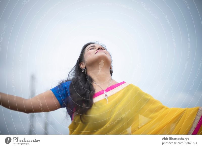 Young and beautiful Indian Bengali brunette woman in Indian traditional yellow sari and blue blouse is standing while spreading her arms on rooftop under blue sky with clouds. Indian lifestyle