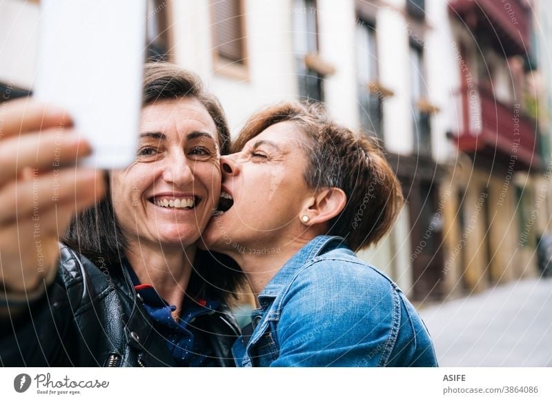 Happy middle aged lesbian couple taking a funny selfie LGBTQ gay 40 50 laughing smartphone self portrait smile embrace hugging holding biting homosexual women