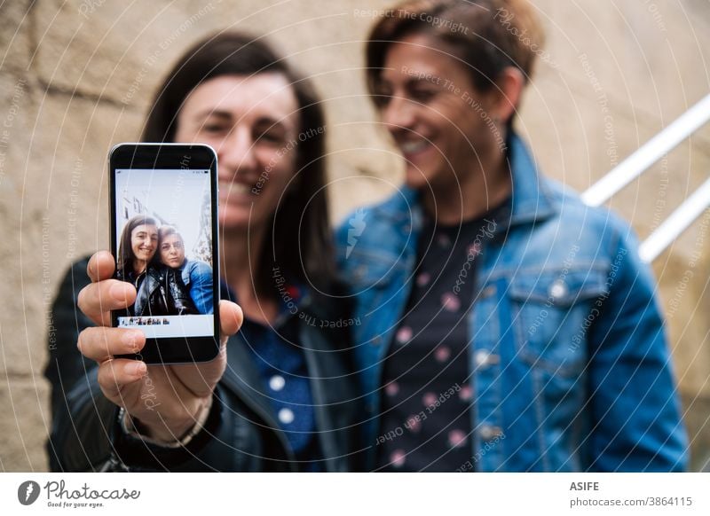 Happy middle aged lesbian couple showing a selfie in the smart phone LGBTQ gay 40 50 laughing smartphone self portrait smile screen embrace hugging holding