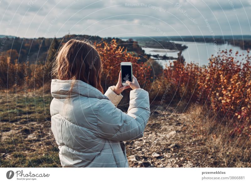 Back view of young woman taking photos of landscape with smartphone during trip on autumn sunny day take selfie outdoors taking selfie destination hiking