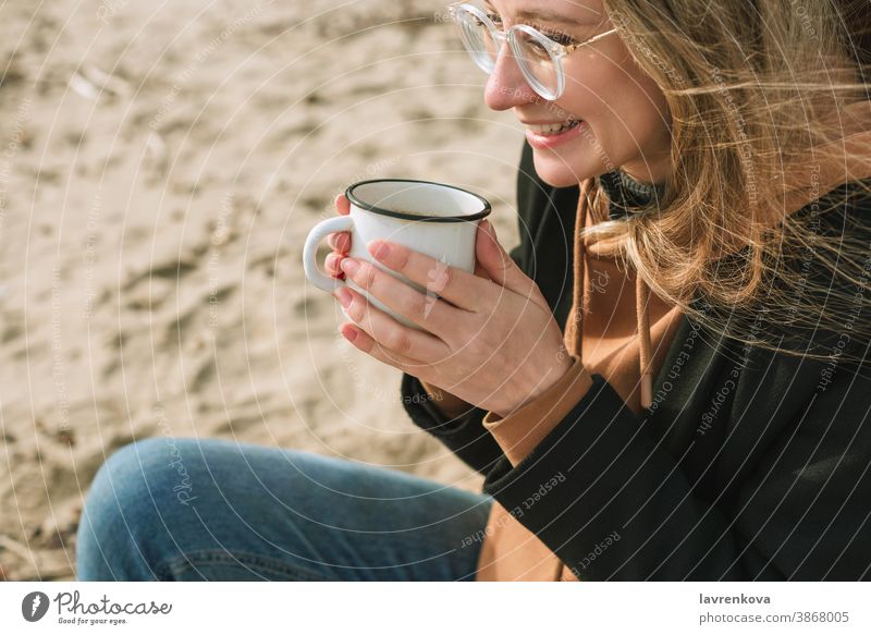 Portait of young adult female with enamel mug with hot drink sitting on a beach woman cup autumn winter happy tea lifestyle smiling morning cheerful seasonal