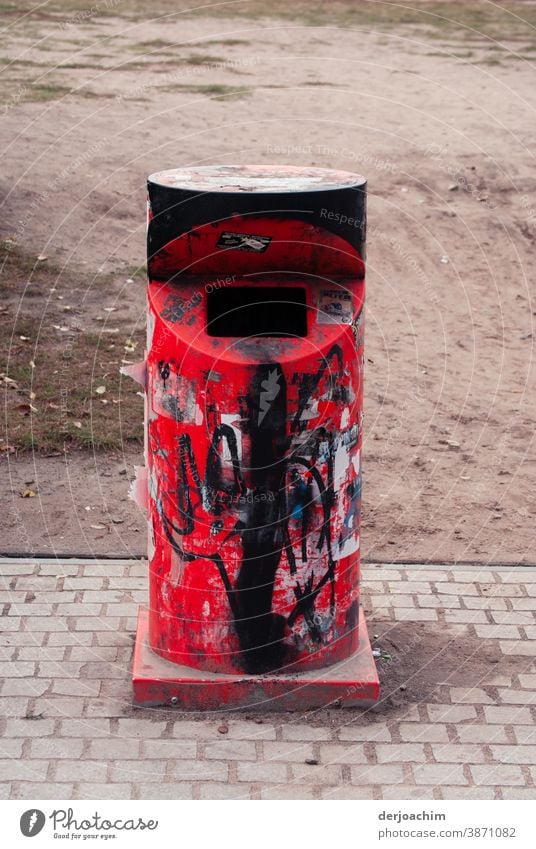 A beautiful red iron litter bin with front throw-in.  Somewhat smudged with black paint, standing lonely and abandoned on the sidewalk. The word NO can still be seen.