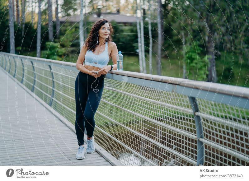 Serious thoughtful woman with spoty body, dressed in cropped top and leggings, poses at bridge looks pensively away, listens music in earphones. People, healthy lifestyle and fitness concept