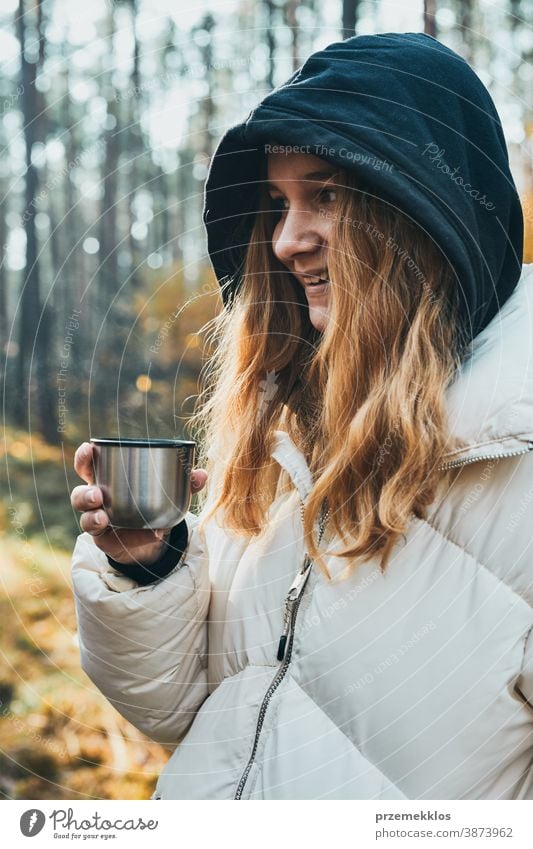 Woman in a hood having break during autumn trip holding cup with hot drink from thermos flask on autumn cold day active activity adventure coffee destination